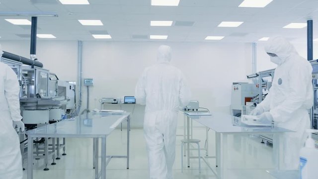 Scientist in Protective Suit Carries Case of Finished Product Through Laboratory. Workers in the Facility with Modern Industrial Machinery. 