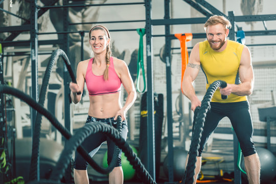 Low-angle view of a strong and competitive couple smiling while exercising together with battle ropes during functional training at the gym
