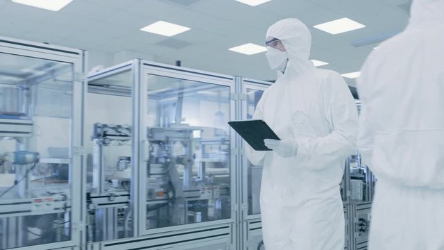Quality Control Check: Scientist Using Digital Tablet Computer and wearing Protective Suit walks through Manufacturing Laboratory. Product Manufacturing: Pharmaceutics, Semiconductors, Biotechnology.