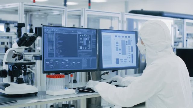 In Laboratory Over the Shoulder View of Scientist in Protective Clothes Doing Research on a Personal Computer. Modern Manufactory Producing Semiconductors and Pharmaceutical Items.