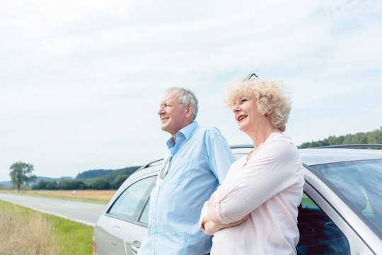 Low-angle View Portrait Of Two Senior People Smiling And Looking Away With Confidence And Positive Attitude While Leaning On Their Car