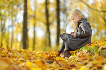 Cute little girl sketching outside on beautiful autumn day. Happy child playing in autumn park. Kid drawing with colourful pencils.