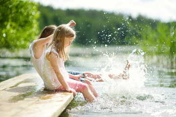 Two cute little girls sitting on a wooden platform by the river or lake dipping their feet in the water on warm summer day - obrazy, fototapety, plakaty