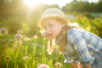 Cute little girl wearing straw hat admiring tall purple garlic flowers on sunny summer day. Child...