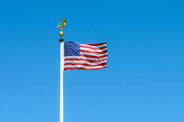 The flag of the United States of America blowing in the wind at full-mast on a white pole topped with a golden eagle on ball against blue sky.