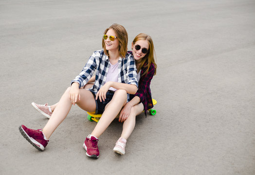 Two smiling female friends having fun riding yellow longboard on the street. Friendship concept
