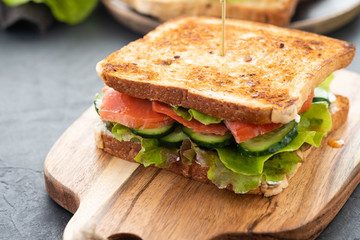 Sandwich with white bread toasts, red fish salmon, fresh green leaves of salad and sliced cucumber on a wooden board.