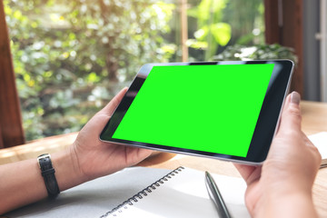 Mockup image of a woman's hands holding black tablet pc with white blank screen with notebook and coffee cup on table