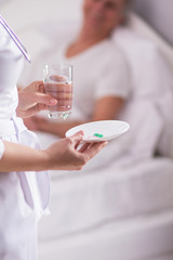 Cropped picture of nurse holding midicine on plate. Female doctor in white coat handing pills and glass of water to patient in bed.