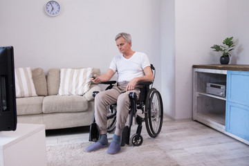 Fototapeta na wymiar Upset disabled man watching TV. Senior man with gray hair sitting in wheelchair in middle of living room witht tv remote in his hand.