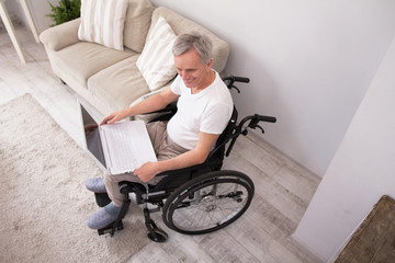 Fototapeta na wymiar Disabled aged man sitting in wheelchair using laptop. Top view of old man smiling while sitting in his living room in wheelchair and websearching via computer.