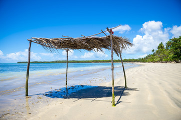 Rustic palm frond and tree branch palapa umbrella waiting to shade visitors to the shallow waters on a remote palm fringed tropical beach Brazil
