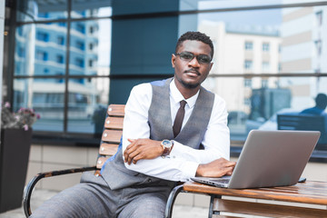 Fototapeta na wymiar african businessman in suit and glasses is sitting at a table with a Laptop, a cup of coffee against a background of glass buildings