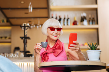 Favourite drink. Positive nice woman drinking espresso while looking at her smartphone screen