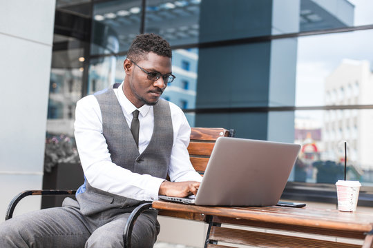 african businessman in suit and glasses is sitting at a table with a Laptop, a cup of coffee against a background of glass buildings