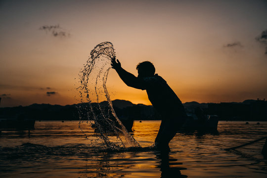 .Young man with funny and relaxed attitude, playing at the seashore throwing water towards the sky at sunset. Lifestyle.