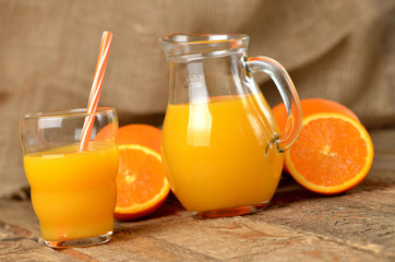 Naklejka na ściany i meble Glass full of orange juice, carafe with fresh juice, straw and fresh oranges in the background on wooden table