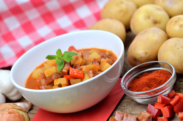 Potato goulash with sausage in white bowl, pepper, onion and garlic on wooden table