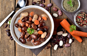 Close-up of full bowl of hot bean soup with large beans on cutting board, carrots, parsley, marjoram, spoon, towel in background - top view