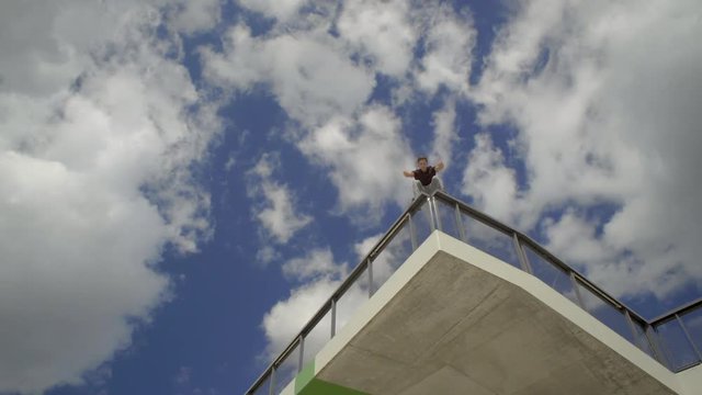 Low and travelling shot of a young parcour runner jumping acrobatic on a railing up high and looking into the distance. 