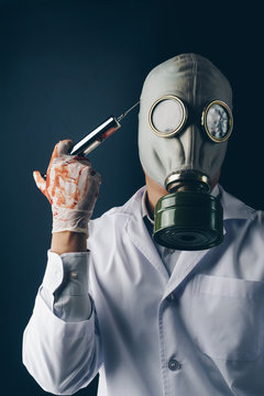 Creepy medical experiment concept, a scary doctor in gas mask wearing bloody gloves with a big stainless steel syringe on dark background