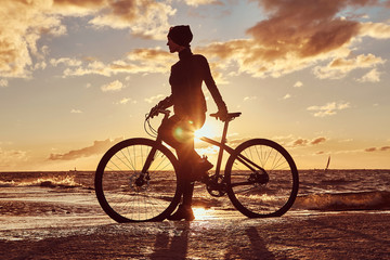 Female cyclist standing with her bicycle and enjoying the sunset on the sea coast.