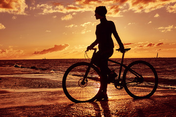 Obraz premium Silhouette of a girl with a bicycle on a sea coast on a sunset background.
