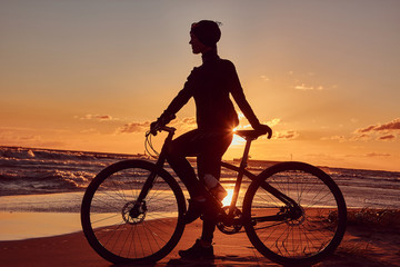 Female cyclist standing with her bicycle and enjoying the sunset on the sea coast.