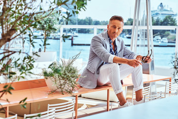 Successful stylish man dressed in modern elegant clothes sitting on table at outdoor cafe against the background of city wharf.
