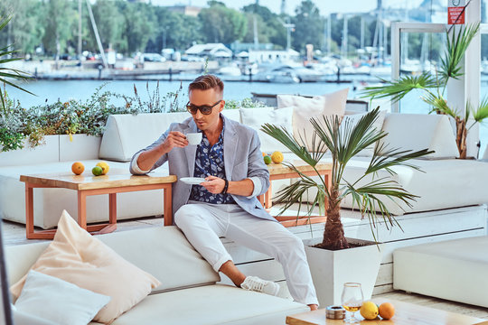 Fashionable Successful Man With Stylish Hair Dressed In Modern Elegant Clothes Holds A Cup Of Coffee While Sitting On A Sofa At Outdoor Cafe Against The Background Of The City Wharf.