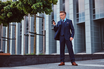 Portrait of a confident stylish businessman dressed in an elegant suit using a smartphone while standing outdoors against skyscraper background.