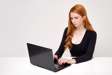 portrait of a beautiful girl with red hair on a white background sitting at the table and working behind a laptop.
