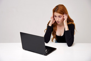 portrait of a beautiful girl with red hair on a white background sitting at the table and working behind a laptop.