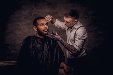 Old-fashioned professional tattooed hairdresser does a haircut to an African American client. Isolated on dark textured background.
