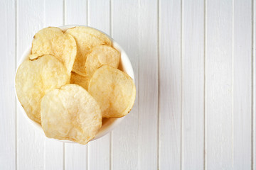 Delicious golden potato chips in a white bowl on a white wooden background.