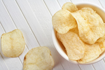Delicious golden potato chips in a white bowl on a white wooden background.