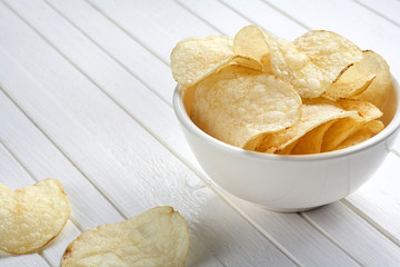Delicious golden potato chips in a white bowl on a white wooden background.