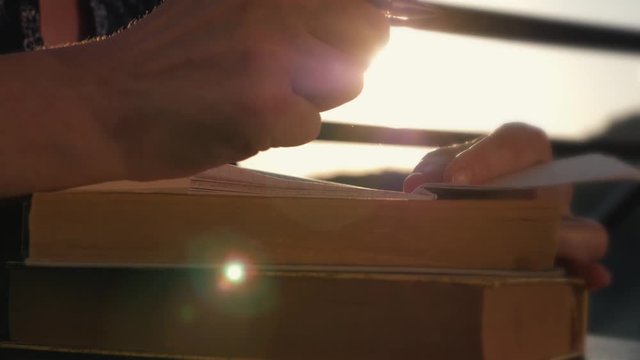 Woman's hand close-up, writing a note or letter to a notebook with a pen on a sunny day at sunset. A stack of scientific books, preparation for exams. Filling out the report