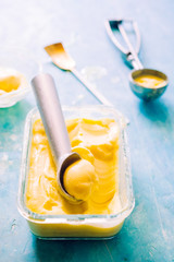 Mango ice cream on a glass container with icecream utensil, on a blue board, frontal view with natural back light