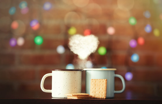 two hot cups of coffee with heart shape steam and fairy lights on background. Christmas season