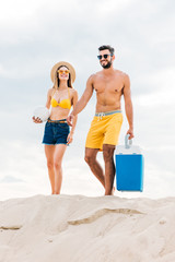 beautiful young couple in beach clothes with volleyball ball and portable fridge walking by sand