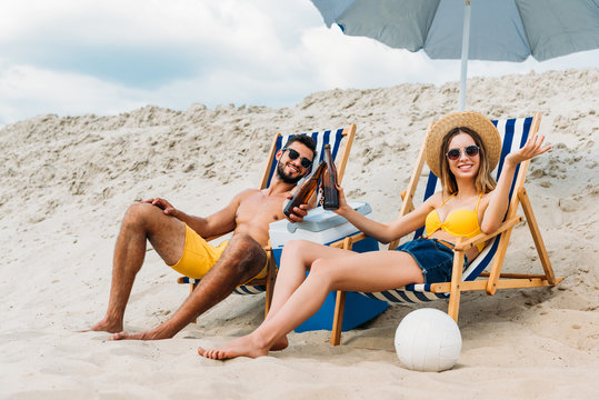 beautiful young couple with beer bottles relaxing in sun loungers on sandy beach - Powered by Adobe