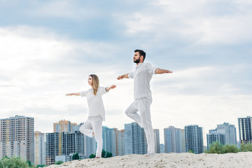 Fototapeta na wymiar beautiful young couple practicing yoga in tree pose (Vrksasana) on sandy dune with city buildings on background