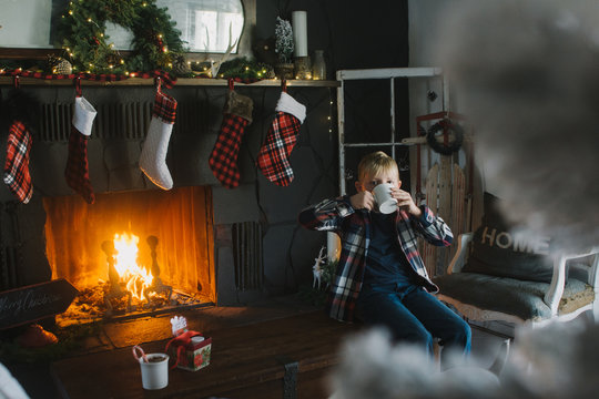 boy sitting on hearth decorated for Christmas drinks cup of coco