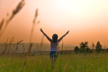 Fototapeta na wymiar Girl in the field on a sunset background. Woman in the meadow with arms raised.