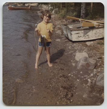 A young boy stands in fron of a rowboat on the shore of a lake h