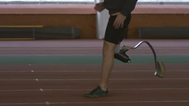 Tilt up of adaptive sportsman with prosthetic leg doing running exercise along track of indoor stadium