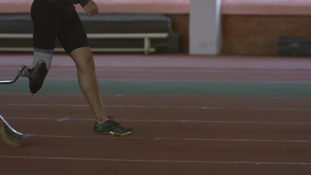 Tilt up of bearded disabled sportsman with prosthetic running leg walking along track in indoor stadium