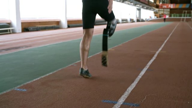 Tracking with rear view of unrecognizable sportsman with prosthetic fitness leg warming up before running practice on track