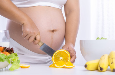 Obraz na płótnie Canvas Closed up on young pregnant with beautiful skin holding a knife slice a fresh orange with vegetables salad bowl and bananas on table. Healthy and fresh food for pregnant.
